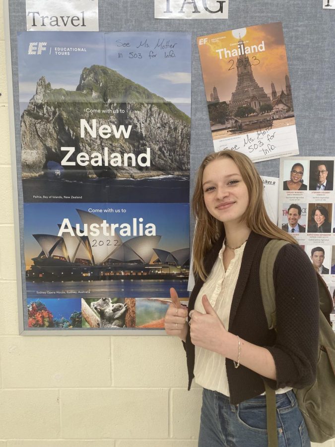 Sophomore Lily Hernandez standing in front of travel posters with her thumbs up. 